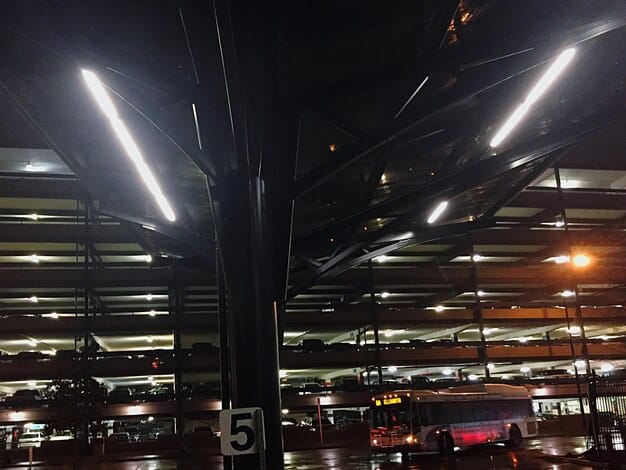A row of energy-efficient LED lights illuminating a warehouse, with workers in the background operating machinery. The lights are bright and evenly distributed.